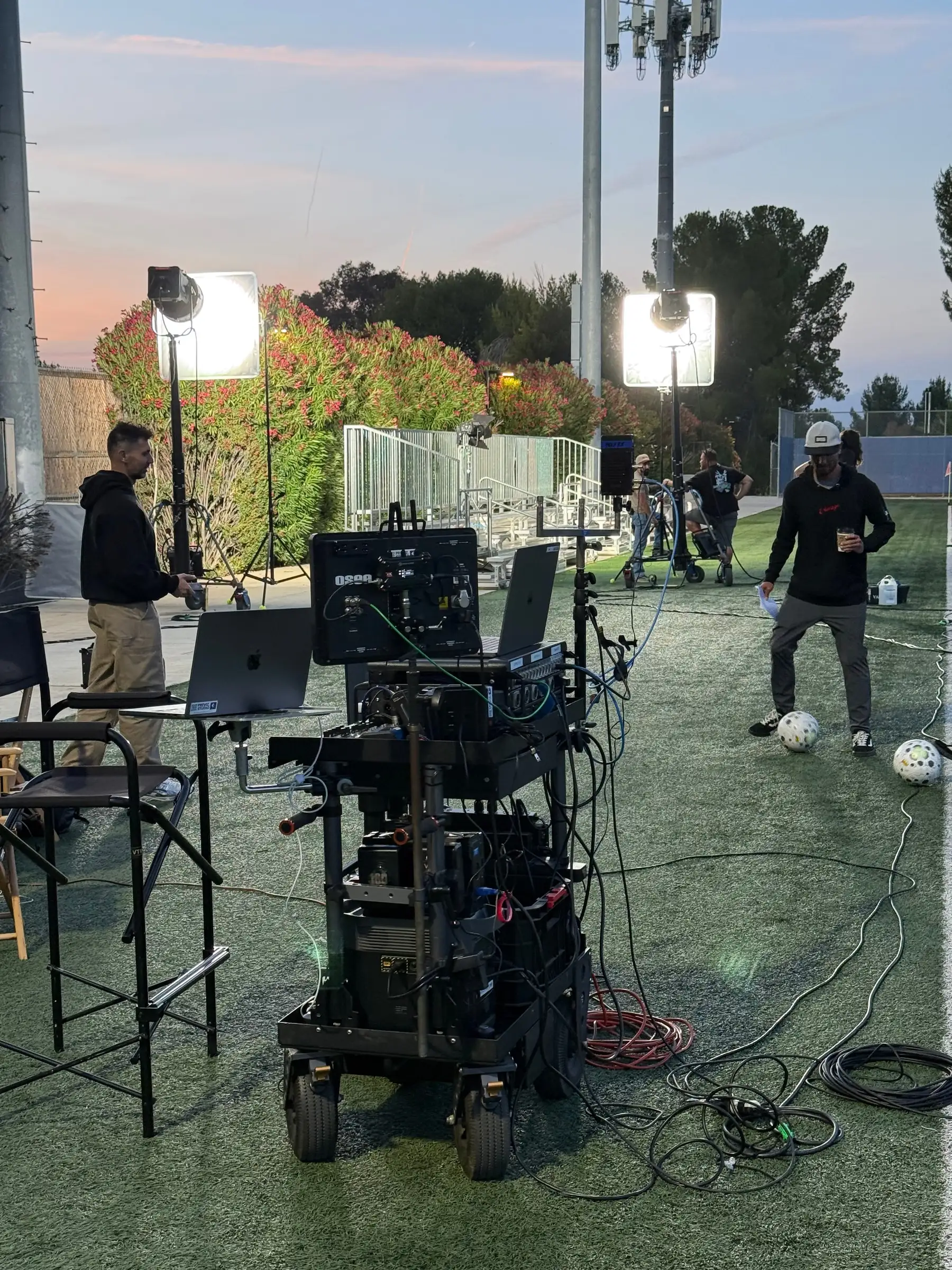 Video-assist cart set up at the edge of the playing area on a dusk exterior, lighting and crew working behind it.