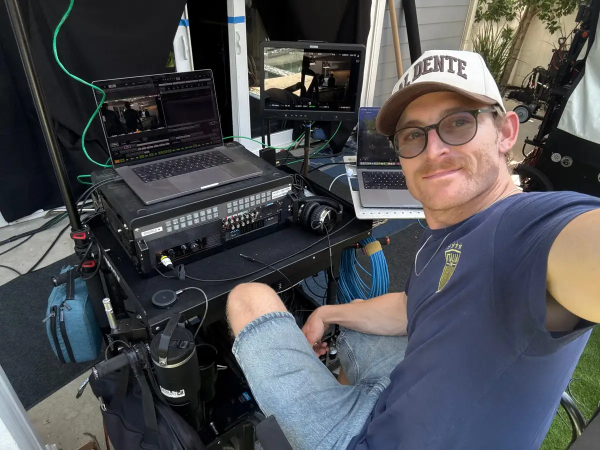 Morgan Spadone at the video-assist position on location, QTAKE running on the laptop beside the router, headphones for production sound, and a camera monitor alongside.